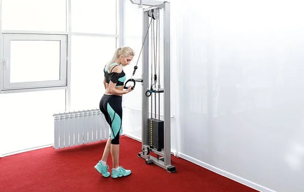 Die athletisch gebräunte Blondine ist im Fitnessstudio. Übungen auf dem Simulator zum Pumpen der Hände im sexy Trainingsanzug. das Konzept eines gesunden Lebensstils. — Stockfoto