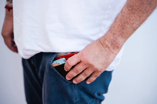 Bir adam yan yan duruyor ve pantolonunun cebine para dolu kahverengi deri bir cüzdan tıkıştırıyor. Beyaz kısa kollu tişörtü ve mavi pantolonu olan sıradan bir kıyafet giyiyor..