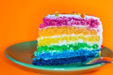 Beyaz kremalı bir dilim Gökkuşağı Pastası. Küçük yeşil bir tabakta servis edilir ve bir kaşık vardır. Tabağın üzerinde duran kekin renkleri yansıtılır. Arka plan turuncu.