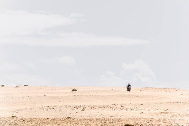 Güneşli bir sabahta mavi gökyüzü ve bulutlarla Fas çölünde motosikletle gezen iki kişi. Boşluğu Kopyala