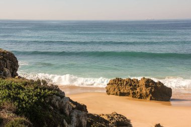 Yosun kayaları olan bir sahil. Kayalardan biri bir dalga almak üzere. Kumsalın kumu temiz ve pürüzsüzdür. Denizin güzel mavi bir rengi var.