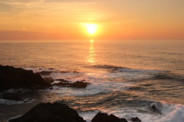 Portekiz 'deki bir uçurumdan portakal rengi bir gün batımı. Güneşin yansıması deniz suyuna güzel bir şekilde yansımıştır. Öfkeli su, sudaki kayalara öfkeyle çarpar. Ufuktaki güneş görüntünün merkezindedir..