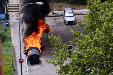 Bir itfaiyeci, konut alanındaki bazı çöp konteynerlerinde çıkan yangının üzerine su döktüğü hortumu tutar. İtfaiyeci neredeyse büyük bir ağacın dallarıyla kaplıdır.