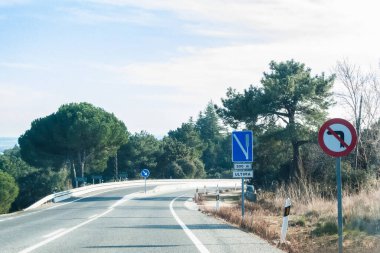 Dağlarda keskin bir virajla iki yönlü yol. Yuvarlak kırmızı tabela bu bölgede sola dönmenin yasak olduğunu gösterir. Arkasında başka bir dikdörtgen, mavi ve dikey işaret 500 metrelik bir acil fren bölgesi olduğunu gösteriyor.