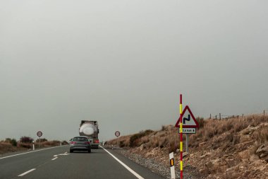 Bir kamyonun arkasında bir araba beklemek zorunda kalacak, çünkü bir dikey geçiş levhasına rastlayacak. Asfaltta gelen şeritten ayrılman gerektiğini gösteren bir ok var. Başka bir tehlike işareti daha var! 