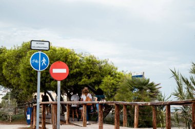 Görüntü, ahşap çitli bir yol boyunca yürüyen çok sayıda yayayı (tanınmaz) gösteriyor. Yolun girişinde bisikletçiler ve yayalar için önceliği belirten dikey bir işaret ve yasaklayan başka bir araç var.