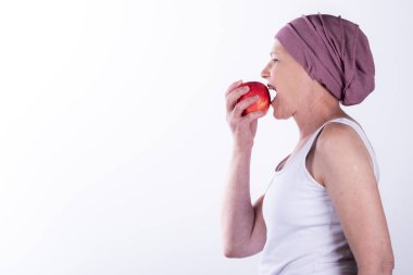 Olgun bir kadın pembe bir başörtüsü ve beyaz bir atkı giyip kırmızı bir elma yiyor. Kanser tedavisi sırasında sağlıklı yaşam tarzını, beslenmeyi ve sağlığını simgeliyor. Beyaz arkaplanda izole.