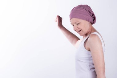 Kutlamak için yumruğunu kaldıran pembe başörtülü neşeli kadın zaferi, güçlenmeyi ve kanserden sonra umut etmeyi simgeliyor. Beyaz arka planda stüdyo çekimi. Reklamcılık, basın makaleleri ya da tıp çalışmaları konsepti. Boşluğu kopyala