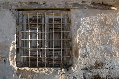Parmaklıklardan yapılmış eski bir duvar penceresi ve kötü bitirilmiş çimento yapısı. Birkaç kusuru olmasına rağmen hepsi beyaza boyanmış. Sabah ışığı yandan içeri giriyor..