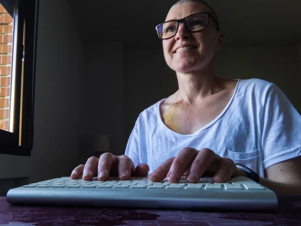 Odasından bilgisayarını uzaktan kontrol eden bir kadın. Eller klavyede dinlenirken yakın plan. Ekrana bakarken gülümsüyor. Kalın bir gözlük, akıllı bir bant, beyaz bir tişört giyiyor ve kemoterapi yüzünden kel. 