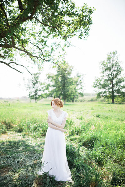 Red-haired bride in white dress embracing herself while standing on grass with eyes closed