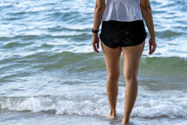 Plajda duran ve denize bakan beyaz gömlekli genç bir kadının bacakları. Asyalı kızın bacakları dinleniyor ve tatilde huzurun tadını çıkarıyor..