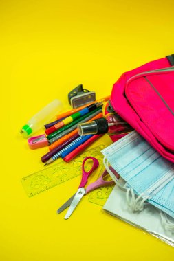 Okul malzemeleri kalem makası silgi cetvel suluboya maske dezenfektanı sarı arka plandaki pembe bir sırt çantasının içinde. Kopya alanı ile covid salgını sırasında okul kavramına geri dön