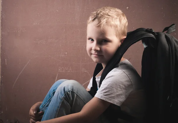 Petit garçon avec un grand sac à dos noir, un gars aux cheveux blonds posant sur un tableau noir , — Photo