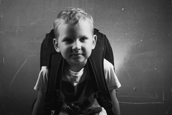 Little boy with a big black backpack, a guy with blond hair posing at a chalkboard, — Stock Photo, Image