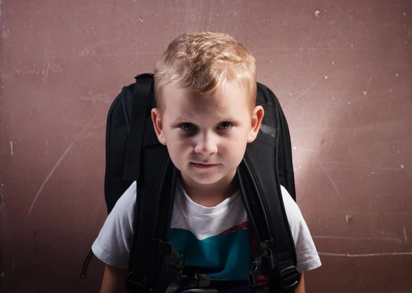 Petit garçon avec un grand sac à dos noir, un gars aux cheveux blonds posant sur un tableau noir , — Photo