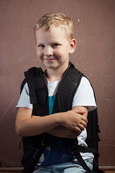 Little boy with a big black backpack, a guy with blond hair posing at a chalkboard, — Stock Photo, Image