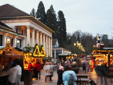 Akşam Baden-Baden Noel market sahnesi aydınlatılmış ahşap tezgahlar, kalabalık ve klasik kumarhane binası.