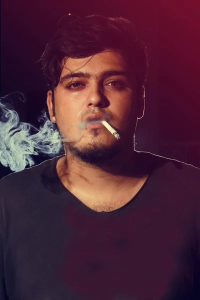 Hombre joven fumando cigarrillo en el fondo oscuro mirando en vino — Foto de Stock