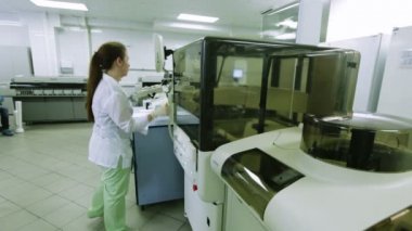 Genç kadın bilim adamı önlük ve eldiven lastik uygun boks bir robot kol ile ve hazır örnekler tepsi ile alır. Tıp