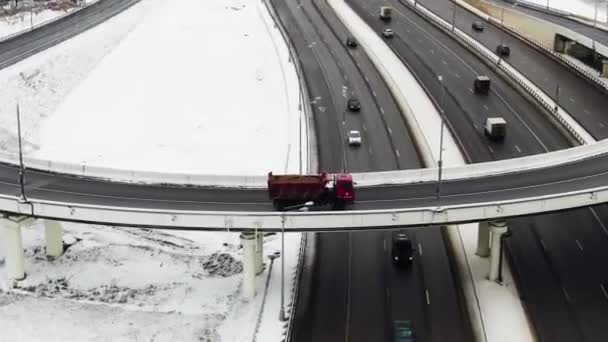 Utsikt över den stora motorvägen i skogen på vintern. En stad i fjärran. En röd lastbil kör genom ett utbyte — Stockvideo