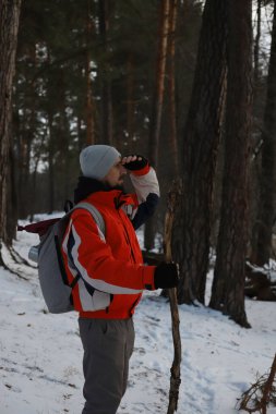 Kışın ormanda yürüyüş yapan bir adam. Gri şapkalı, turuncu ceketli ve gri sırt çantalı bir adam..