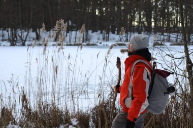 Kışın ormanda yürüyüş yapan bir adam. Gri şapkalı, turuncu ceketli ve gri sırt çantalı bir adam..