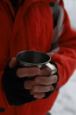 Bir kış ormanında, turuncu ceketli bir adam elinde bir teneke bardak tutuyor ve ondan sıcak bir içecek içiyor..