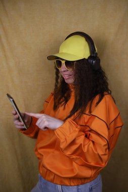 Sarı şapkalı, sarı gözlüklü ve turuncu retro spor ceketli bir kız kulaklıkla müzik dinliyor ve elinde bir cep telefonu tutuyor..