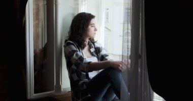 Pencere pervazında oturan ve eski kasaba manzarasının tadını çıkaran genç bir kadın. Evde temiz hava alan rahatlatıcı bir kız silueti.