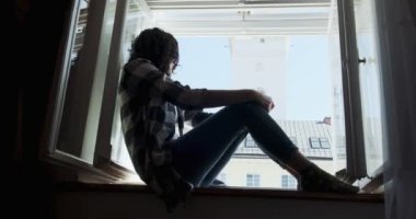 Pencere pervazında oturan ve eski kasaba manzarasının tadını çıkaran genç bir kadın. Evde temiz hava alan rahatlatıcı bir kız silueti.