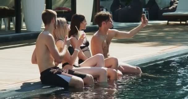 Kelompok teman bersenang-senang di kolam renang. Mengambil foto selfie di smartphone duduk bersama di samping kolam renang. Pesta musim panas dekat kolam renang Stok Video