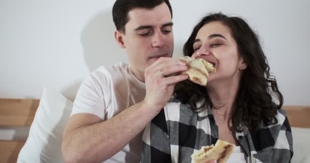 幸せな夫婦と女性は自宅のベッドでピザを食べています。幸せな若い恋人は一緒に過ごす時間を楽しむ — ストック動画