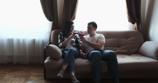 Романтична пара святкує успіх з червоним вином. Успішна сім'я п'є алкоголь у вітальні на дивані. Красивий чоловік приносить келихи вина дружині Ліцензійні Стокові Відео