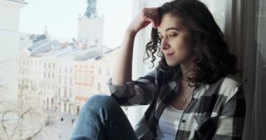 Pencere pervazında oturan ve eski kasaba manzarasının tadını çıkaran genç bir kadın. Evde temiz hava alan rahatlatıcı bir kız silueti.