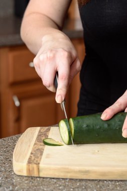 Mutfakta görülebilen eller kesme tahtasındaki sebzeleri bıçakla kesiyor. Bıçak bir salatalığı dilimliyor..
