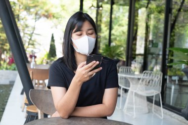 Kafede akıllı telefon kullanan tıbbi maske takan genç bir Asyalı kadın. Modern kafede cep telefonuyla mesaj ya da sosyal medya okuyan Asyalı bir kadın. Yeni normal ve sosyal uzaklık çalışıyor.