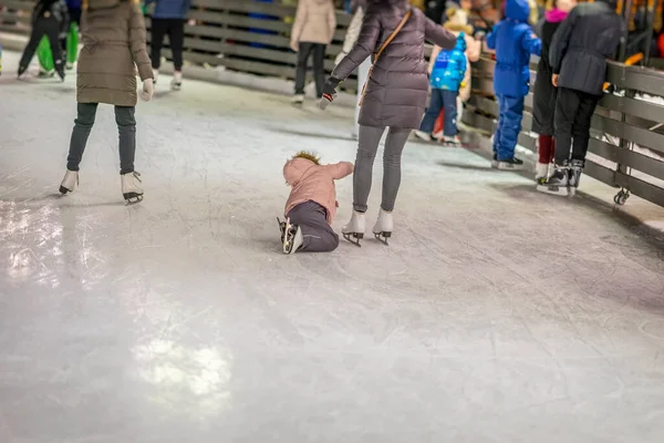 Каток. Мама помогает воспитывать дочь. Маленький ребёнок учится кататься на коньках и падает на лёд. Зимние каникулы в городе на катке — стоковое фото