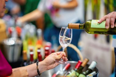 Tat kabinine beyaz şarap dökülüyor. Beyaz şarabın bardağa döküldüğü yakın çekim, şarap tatma etkinliği. Cam ve şişeli eller, bulanık şişeler, arka planda insanlar. Şarap fuarı