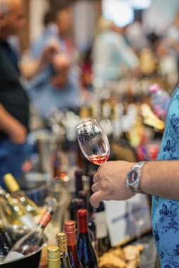 Adamın elinde şarap kadehi, şişe ve insanlarla çevrili şarap tatma etkinliği, fuar atmosferi var. Şarap kültürü kavramı, sosyal hizmet toplantısı.