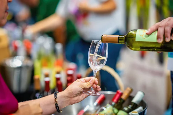 Tat kabinine beyaz şarap dökülüyor. Beyaz şarabın bardağa döküldüğü yakın çekim, şarap tatma etkinliği. Cam ve şişeli eller, bulanık şişeler, arka planda insanlar. Şarap fuarı