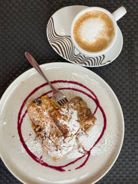 Seramik tabakta, pudra şekeri ve böğürtlen şurubuyla servis edilen bir bardak kapuçino. Sıcak kahve ve mevsimlik lezzetlerle Şükran Günü tatili.