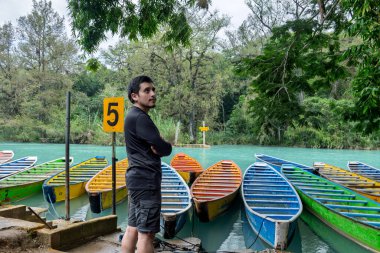 Huasteca Potosina 'daki Cascada del Meco nehrinin kenarındaki renkli teknelerin yanında bir kişi duruyor. Bölge tropikal bitkiler ve ağaçlarla dolu. Sahne canlı ve canlı..