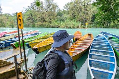 Huasteca Potosina 'daki Cascada del Meco nehrinin kenarındaki renkli teknelerin yanında bir kişi duruyor. Bölge tropikal bitkiler ve ağaçlarla dolu. Sahne canlı ve canlı..