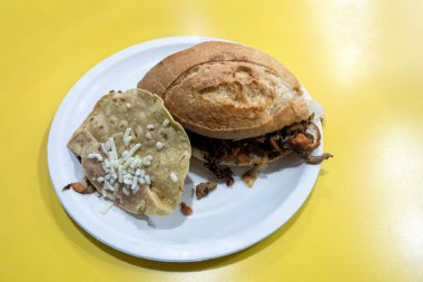 Meksika sandviçi et ve malzemelerle dolu, yanında da Comifa Mexicana 'da peynirli bir tortilla. Ayarda parlak sarı bir masa var..