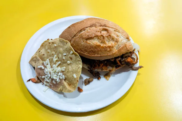 Meksika sandviçi et ve malzemelerle dolu, yanında da Comifa Mexicana 'da peynirli bir tortilla. Ayarda parlak sarı bir masa var..