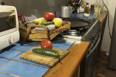 Birçok taze sebze mutfaktaki ahşap bir kesme tahtasının üzerinde oturur. Sepette daha fazla sebze var. Arka planda bir soba ve diğer mutfak malzemeleri var..