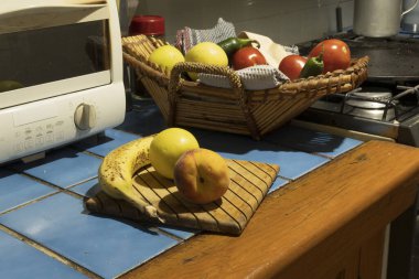 Meyve ve sebzeler mutfak tezgahındaki tahta kesme tahtasına dizilir. Yakınlarda bir sepet daha var. Pencereden gelen ışık, yoğun bir mutfağı gösteriyor..