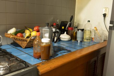 Mutfak tezgahında sebze ve meyve sepeti, yemek aletleri, yağ şişeleri ve kahve makinesi var. Aydınlık ve düzenli bir ortam, yemek hazırlığını vurguluyor..