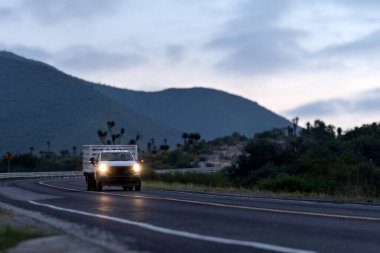 Güneş dağların ardında batarken ve bulutlar akşam gökyüzünü doldururken bir kamyon dolambaçlı bir yolda ilerliyor. Sahne doğayı ve ulaşımı gösteriyor..